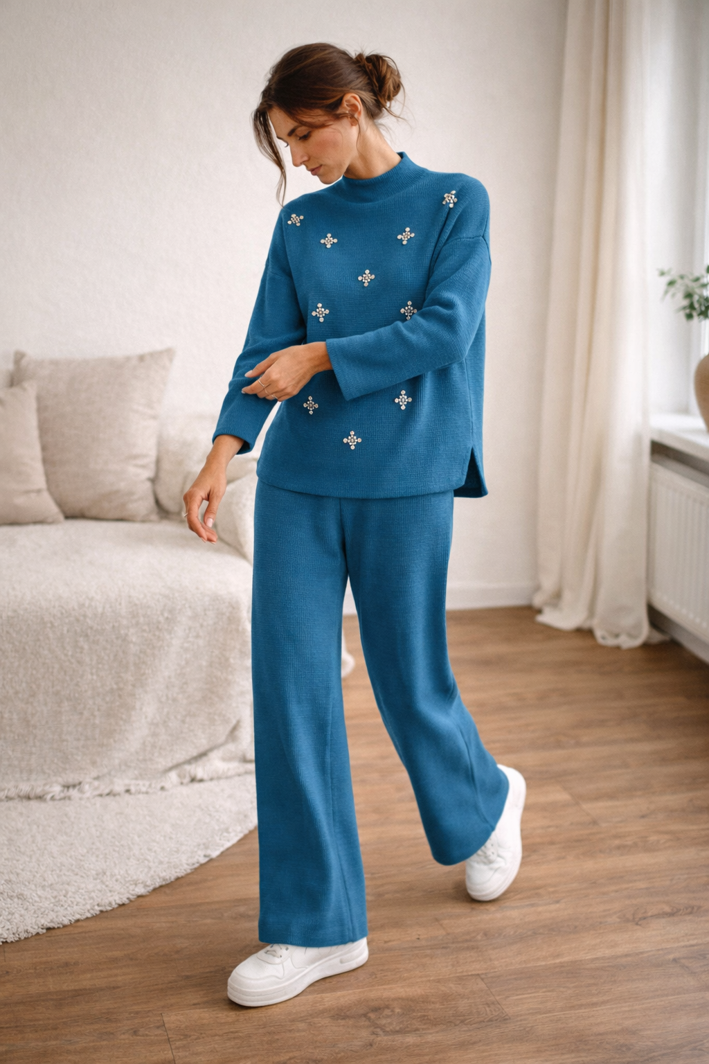 Woman wearing a blue outfit with decorative elements in a room with a couch and curtains.