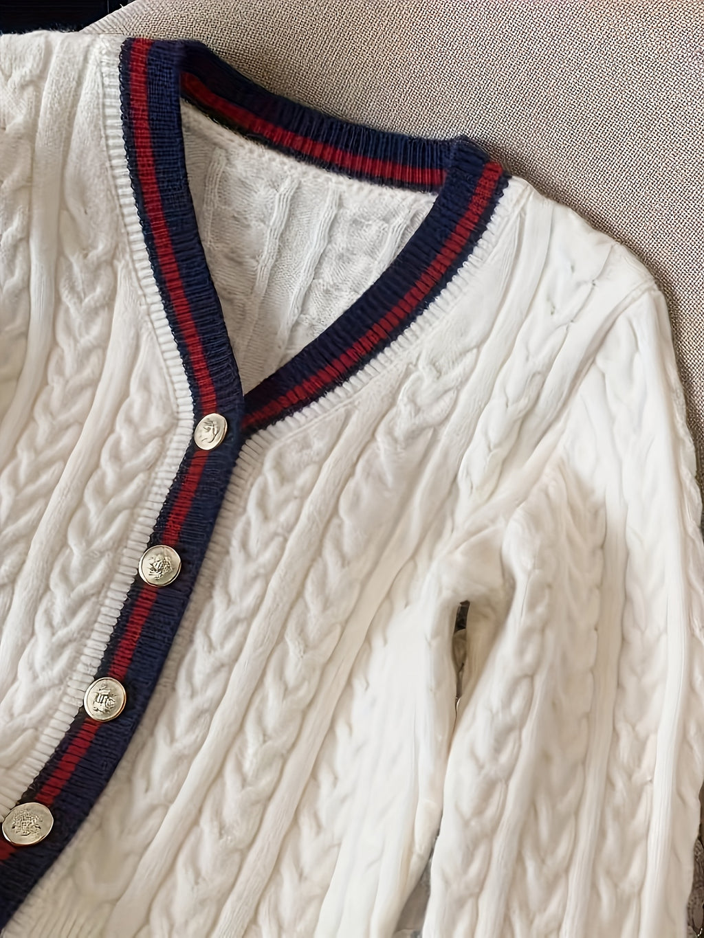 White cable knit cardigan with navy and red trim and gold buttons on a neutral background