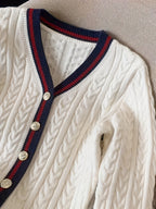 White cable knit cardigan with navy and red trim and gold buttons on a neutral background