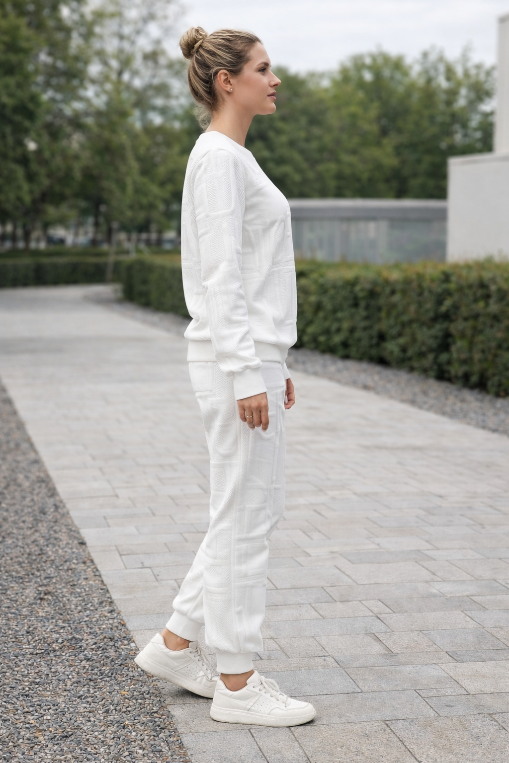 Woman in a white outfit standing on a paved path with greenery in the background