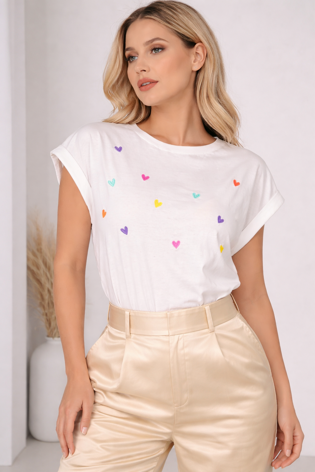 Woman wearing a white t-shirt with colorful heart patterns and beige pants against a white background