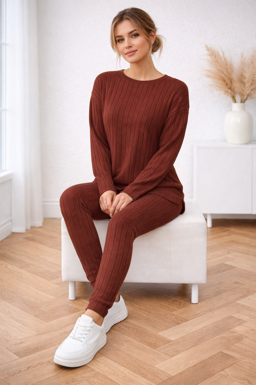 Woman in a brown ribbed outfit sitting on a white stool in a room with wooden flooring.