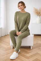 Woman sitting on a white stool wearing a green ribbed sweater and pants set in a room with wooden flooring.