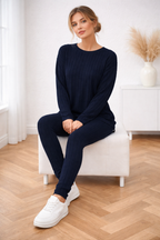 Woman sitting on a white chair wearing a navy blue outfit with white sneakers in a room with wooden flooring.