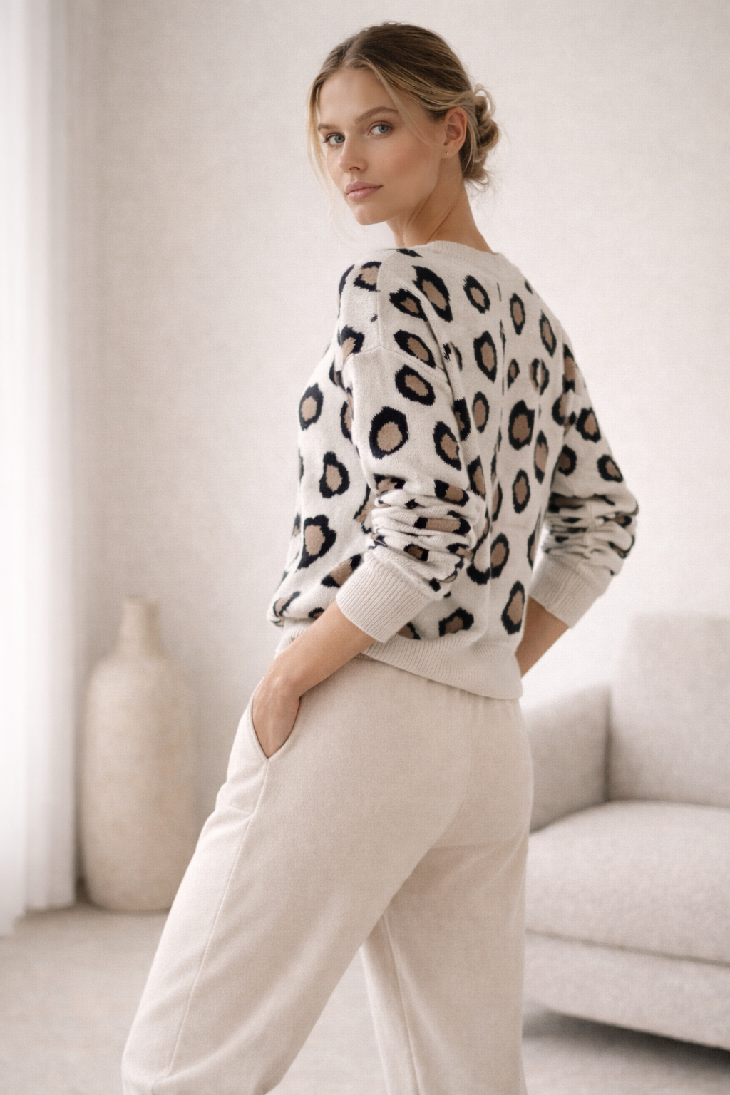 Woman wearing a leopard print sweater and white pants in a minimalistic room.