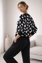 Woman wearing a black sweater with white patterns and black pants in a minimalistic room.