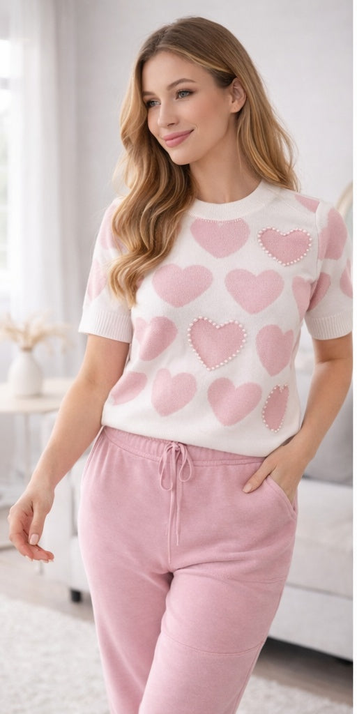 Woman wearing a white sweater with pink heart patterns and pink pants in a bright room.