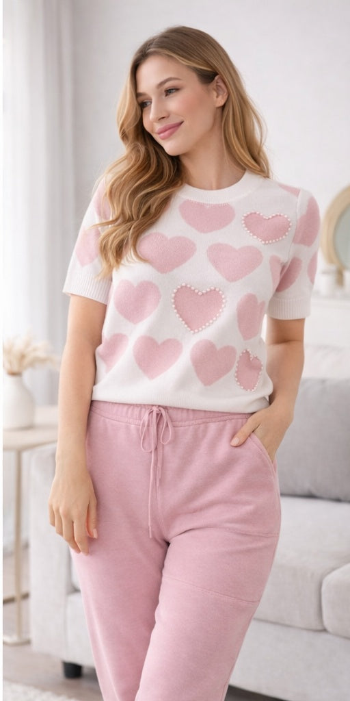 Woman wearing a white sweater with pink heart patterns and pink pants in a living room.