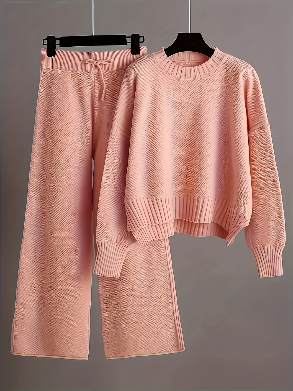 Pink knit sweater and pants set on hangers against a gray background