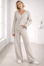 Woman wearing a light gray cable knit sweater and pants in a bright room.