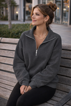 Woman sitting on a bench wearing a gray zip-up sweatshirt.
