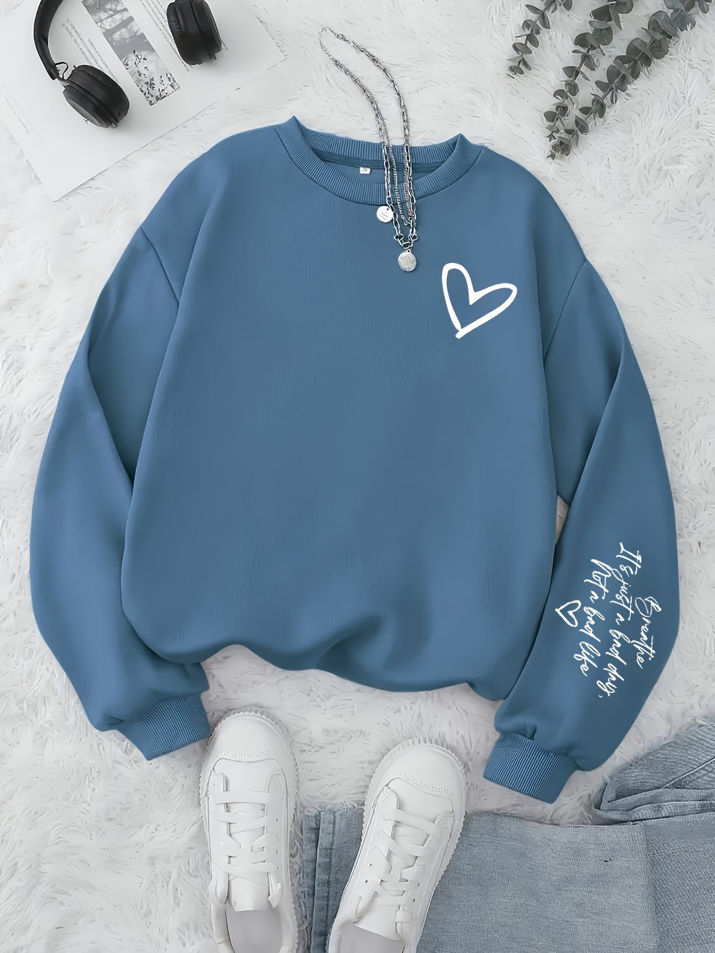 Blue sweatshirt with a heart design on a light gray surface with sneakers and headphones.