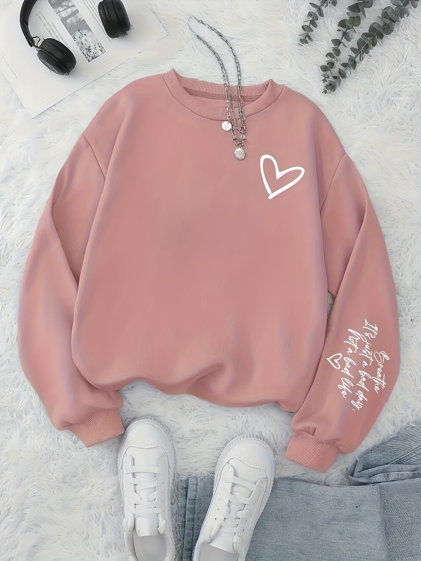 Pink sweatshirt with a heart design on a light gray surface with sneakers and headphones.