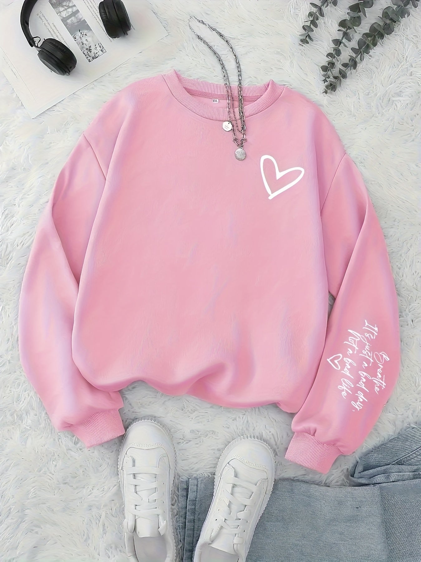 Pink sweatshirt with a heart design on a light gray surface with sneakers and headphones.