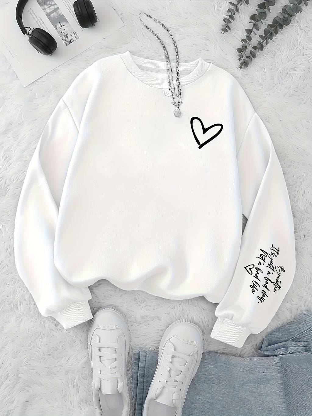 White sweatshirt with black heart design on a light gray surface with sneakers and headphones.