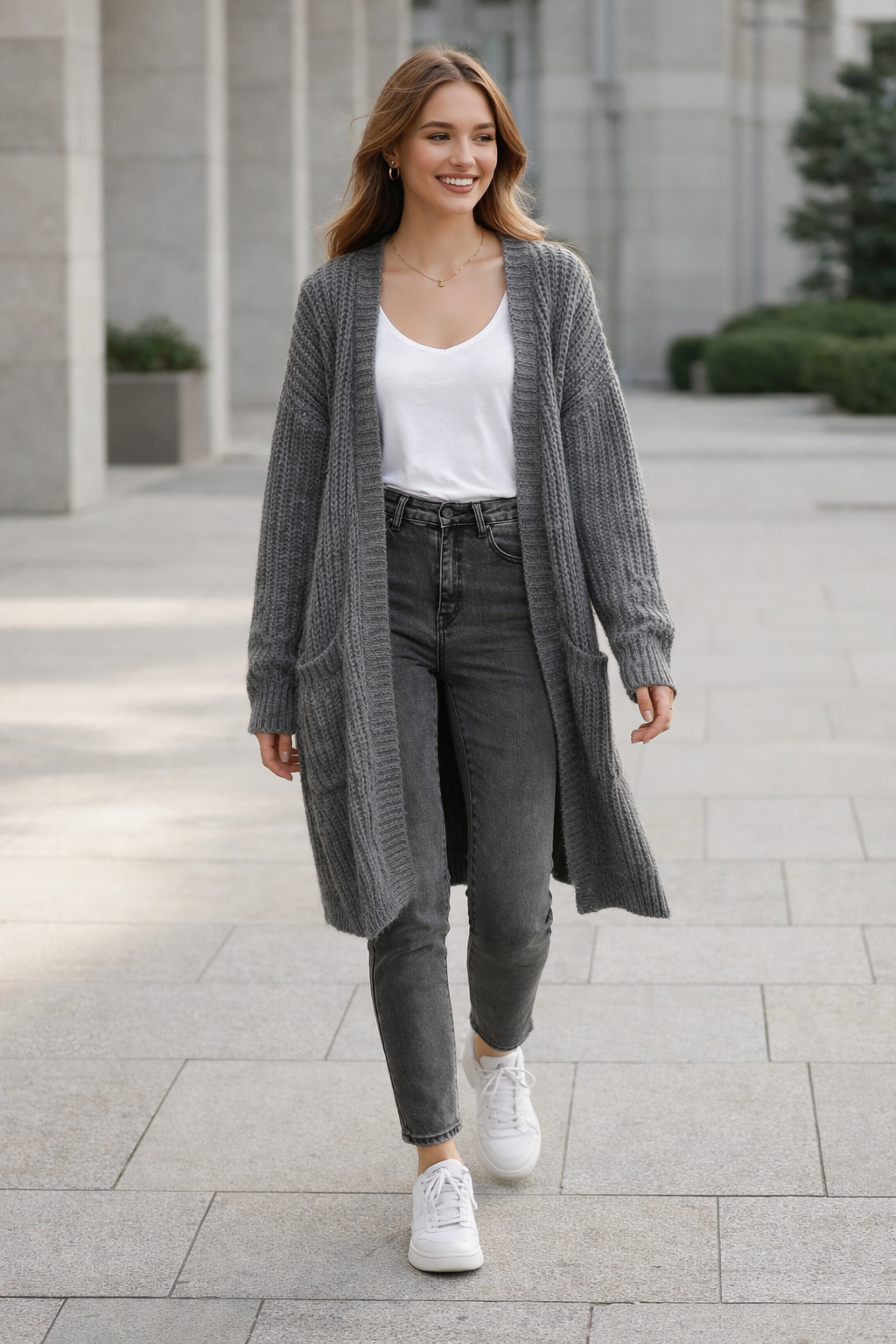 Woman wearing a long gray cardigan, white top, gray pants, and white sneakers on a city street.