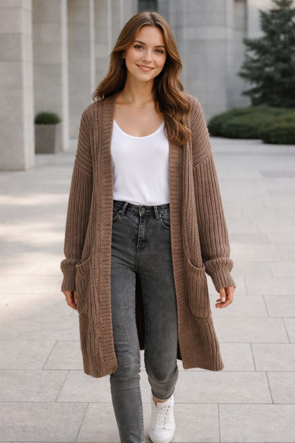 Woman wearing a long brown cardigan, white top, and gray jeans outdoors.