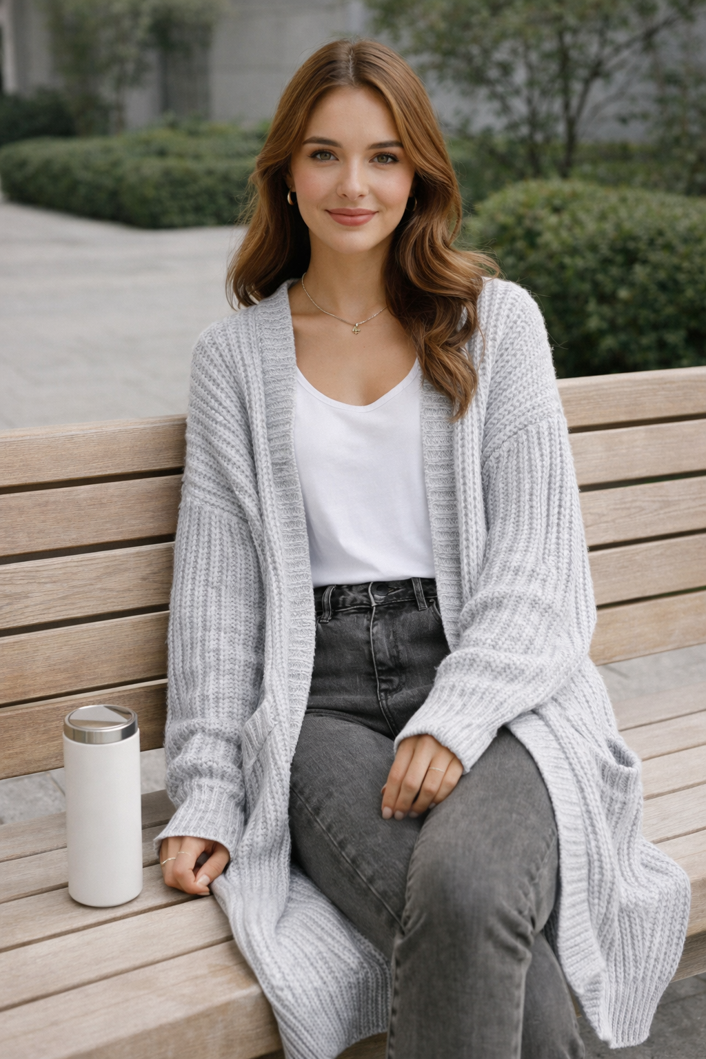 Woman sitting on a wooden bench wearing a light gray cardigan, white shirt, and dark jeans.