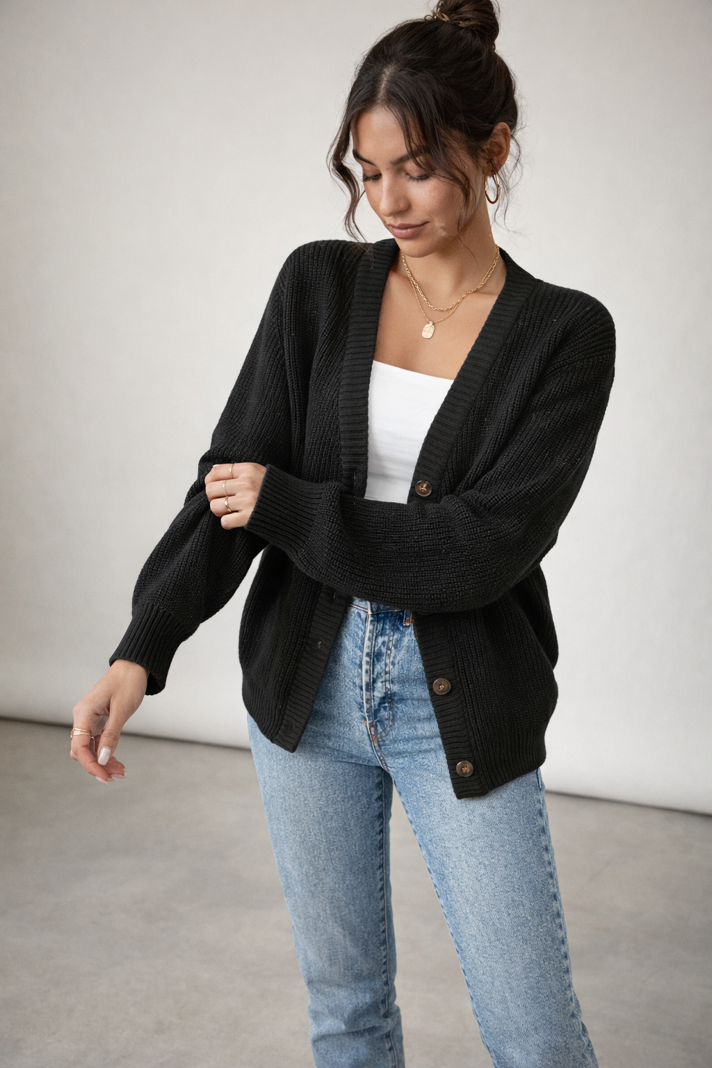 Woman wearing a black cardigan over a white top and blue jeans against a plain background