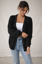 Woman wearing a black cardigan over a white top and blue jeans against a plain background
