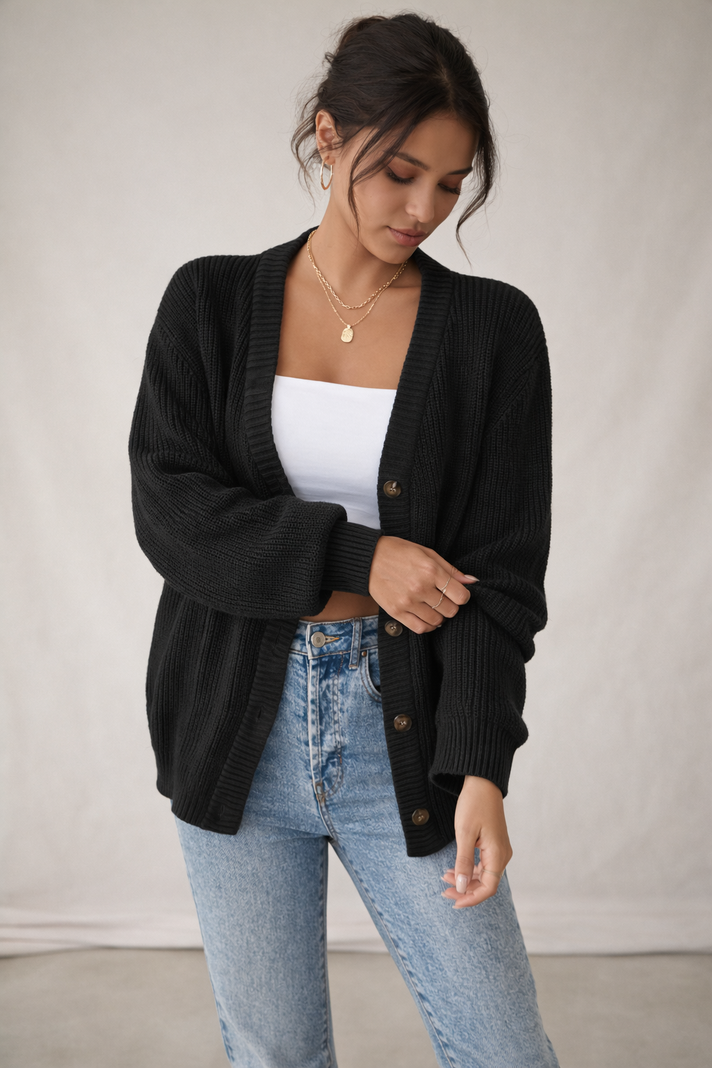 Woman wearing a black cardigan over a white top and blue jeans against a plain background