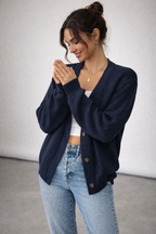 Woman wearing a navy blue cardigan over a white top and blue jeans, standing against a plain background.